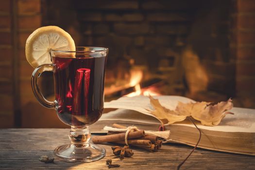 Glass of mulled wine, spices, a book on a table in a cozy room with a burning fireplace.