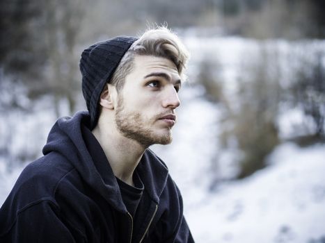 Portrait of young man in hoodie posing outdoor in winter setting with snow all around, looking away to a side.