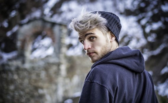 Portrait of young man in hoodie posing outdoor in winter setting with snow all around, looking at camera.
