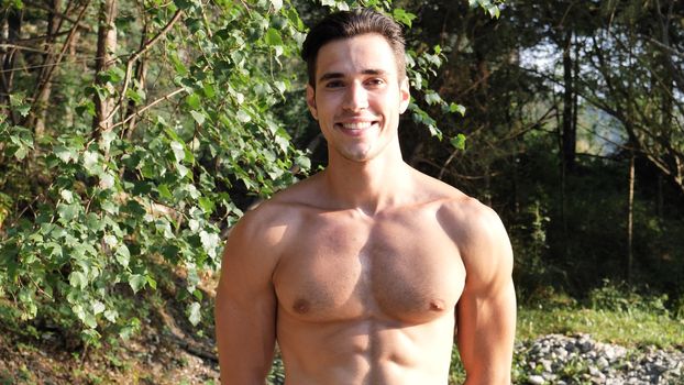 Handsome athletic shirtless young man in nature smiling, in a sunny, peaceful day, looking at camera