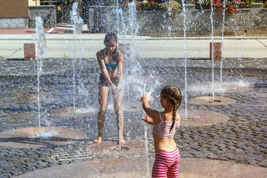 Children on a sunny summer day are poured water from a fountain.Children happily in shallow clean water on of city fountain on warm bright summer day.