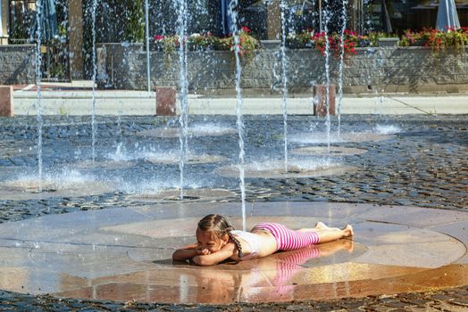 Girl on a sunny summer day are poured water from a fountain.Girl happily in shallow clean water on of city fountain on warm bright summer day.