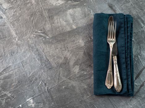 Silver fork and knife on dark green emerald linen napkin over gray textured background table with copy space. Cutlery with copy space, top view or flat lay
