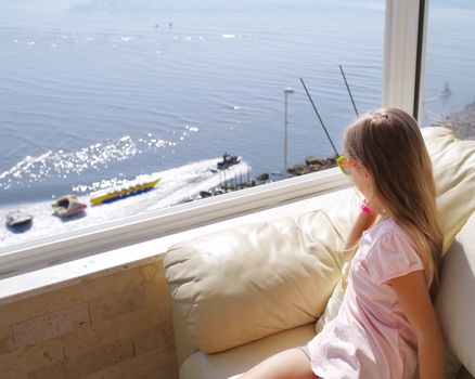 Rear view of young woman looking at sea view from balcony at resort.A little girl sits by the window and looks at the ocean. The view from the window to the sea.