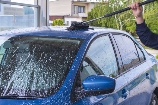 Manual car wash with pressurized water in car wash outside.Using a brush to wash a car on a car washing facility on sunny summer day.Blue car manual wash of with pressurized water in car wash outside.