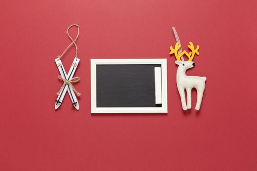 Christmas toys wooden skis, white felt toy deer and small clean chalk board with copy space on dark red background. Festive, New Year concept. Horizontal, flat lay. Minimal style. Top view.