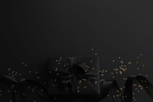 Black friday gift, paper box with silk ribbon bow and golden glitter stars on black paper background with copy space for text, flat lay top view template