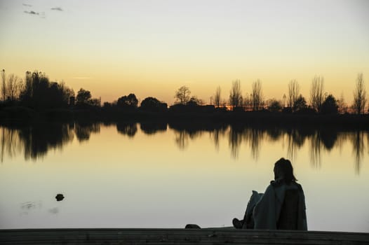 Young boy sitting in front of the lake while watching the sunset with its colors and the sun going down under the lake horizon.