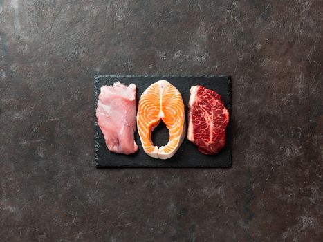 Raw turkey breast fillet, salmon steak and beef ribeye steak on black slate board over dark background. Top view or flat lay. Protein food, keto, carnivore, paleo diet concept. Copy space.