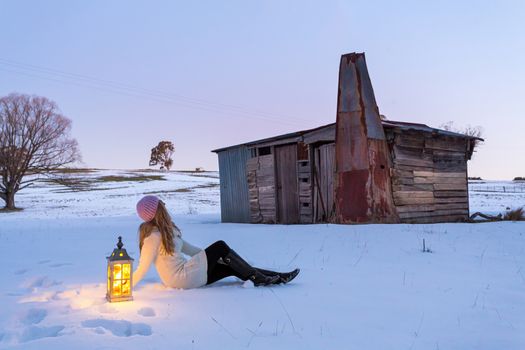 Woman with lantern sitting in a snow covered field.  The day is drawing to a close and the cool dusk colours envelop the landscape.  A rustic stable shed  of mismatched timber and metal nearby