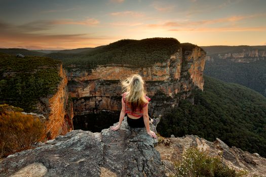 A female sits down on a rock and relaxes to watch the sunset with views of mountains and valleys.  Winds blowing her hair long blond hair every which way