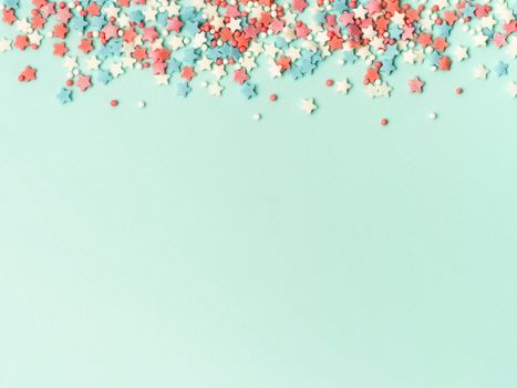 Festive border frame of colorful pastel sprinkles on blue background with copy space bottom. Sugar sprinkle dots and stars, decoration for cake and bakery. Top view or flat lay.