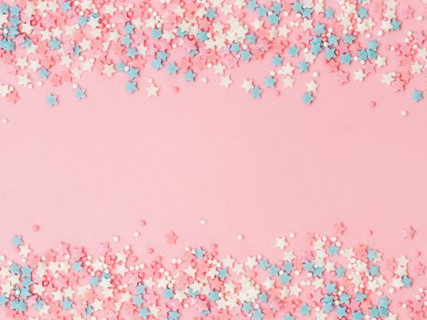Festive border frame of colorful pastel sprinkles on pink background with copy space in center. Sugar sprinkle dots and stars, decoration for cake and bakery. Top view or flat lay