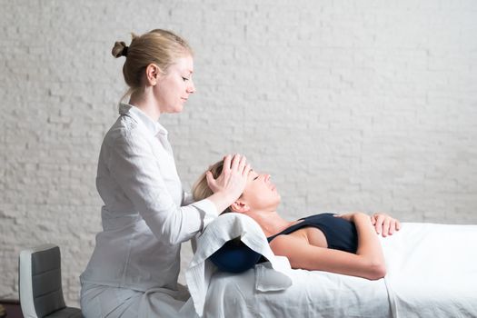 Professional female masseur giving relaxing massage treatment to young female client. Hands of masseuse on forehead of young lady during procedure of spa facial massage.