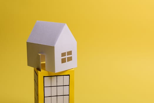 White family paper house over block of flats on yellow background paper. Minimalistic and simple concept, style. Copy space. Vertical orientation.