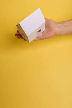 White family paper house in man hand on yellow background paper. Minimalistic style. Copy space. View from above. Horizontal orientation