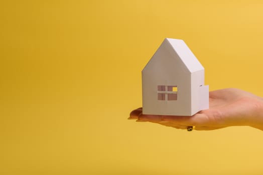 White family paper house in man hand on yellow background paper. Minimalistic style. Copy space. View from above. Horizontal orientation