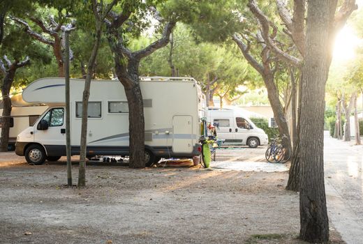 White camper on campsite. Sun Rays in the morning through the trees. Many bikes and caravans in camping.