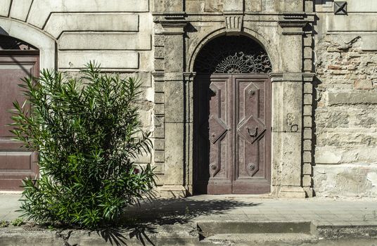Old typical italian wooden door. Italian house. Ancient house facade. Sunlight. Round door arch. Stone build house. Wrought iron door handles. Green decorative flower.