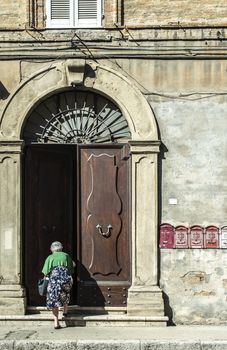 Senior woman enter in the house. Old typical italian wooden door. Italian house. Ancient house facade. Sunlight. Round door arch. Stone build house. Wrought iron door handles.