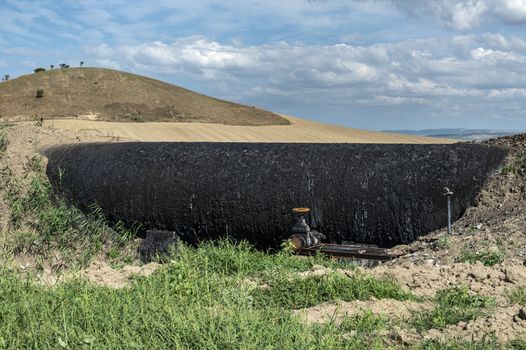 Black Oil pipeline goes out above the ground. Oil-stained pipe. Concept for oil petrol transportation with tubes. Green grass and black color oil pipe with faucet.