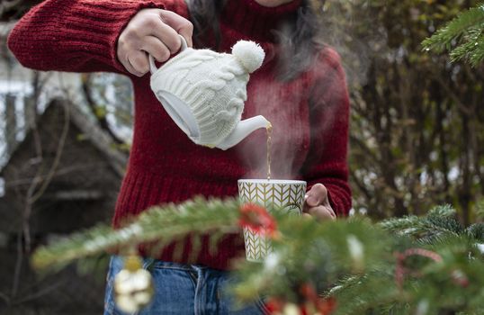 Woman pours tea with a teapot into a teacup. Christmas tree in the foreground. Teacup with Christmas ornaments. Steam comes out of cup of tea. Winter and Christmas concept. Exterior shot.