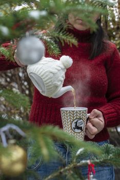 Woman pours tea with a teapot into a teacup. Christmas tree in the foreground. Teacup with Christmas ornaments. Steam comes out of cup of tea. Winter and Christmas concept. Exterior shot.
