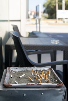 Ashtray with sand and buried cigarettes. Table and chairs. Many cigarette butts.