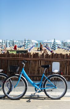 Rental bikes on the beach. Blue bicycles on the street. Many beach umbrellas.