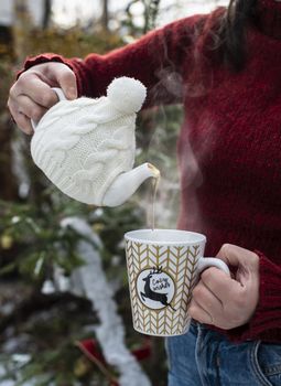 Woman pours tea with a teapot into a teacup. Christmas tree in the foreground. Teacup with Christmas ornaments. Steam comes out of cup of tea. Winter and Christmas concept. Exterior shot.