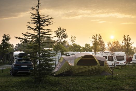 Caravans and tent on green meadow in campsite. Sunrise, rays on campers and tent in the morning. Green grass. Outdoor concept for traveling and res in the nature.