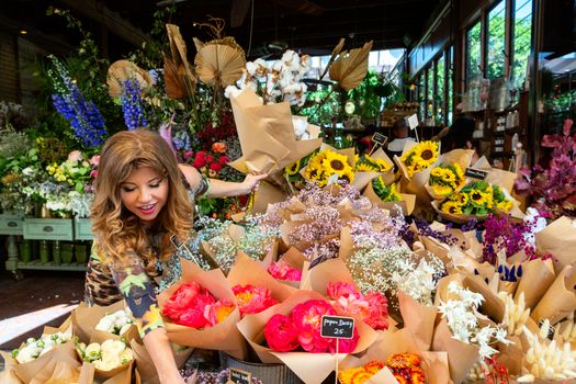 Woman on the footpath outside the florist choosing bunches of flowers on display, behind her the shop is open plan industrial look with rustic features.