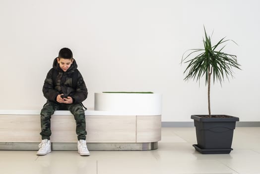 Teenager playing with smartphone in modern commercial center. Bench and flower in a pot. Modern building interior. Technology and communication concept with child in contemporary building interior.