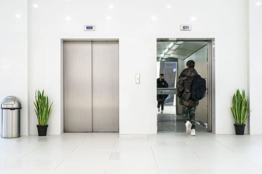 Teenager boy with casual clothes and white sneakers enters in elevator. White contemporary building interior. Flowers in pots and white wall. Metallic elevator doors.