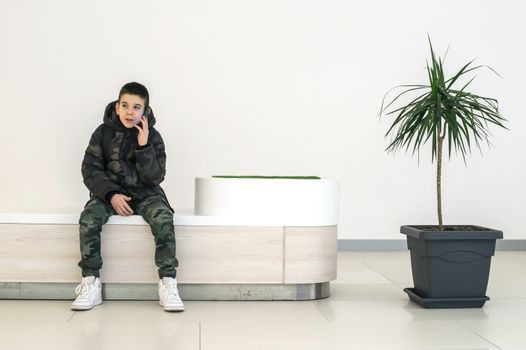 Teenager talking on smartphone in modern commercial center. Bench and flower in a pot. Modern building interior. Technology and communication concept with child in contemporary building interior.