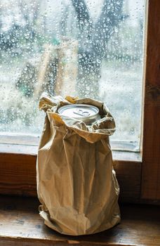 Bottle with alcohol in a brown pack paper placed to the window at rainy day. Wrinkled paper package. Hidden bottle alcohol in paper. Concept for stealthily drinking and alcoholism.