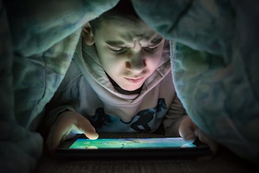 Child watching his tablet in the bed. Illuminated child face from device screen. Boy under the covers hold a tablet. Night time.