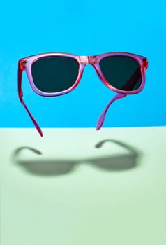 Bright pink colour sun glasses in the air. Floating in the air pink sunglasses on pastel tones blue and green background.  Lower shadow below the glasses. Minimalism summer concept.