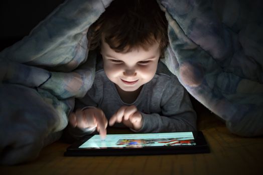 Little girl watching her tablet in the bed. Illuminated smiling child face from device screen. Child dressed with pajamas under the covers hold a tablet. Night time.
