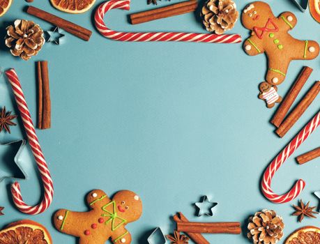 Frame of Christmas gingerbread cookies on blue background with copy space. Pattern of gingerbread men, candy canes, star mold, cinnamon, pine cone, orange. Top view, flat lay.