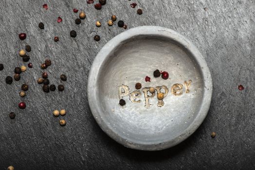 Black pepper in small bowl for spices on dark background. Red, green and black pepper grains close-up and natural light on it. Dark stone background and bowl with text Pepper on the bottom.