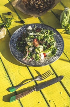 Green salad with arugula, Little Gem, cheese, olives, tomatoes, cucumber, onion and caramelized nuts in old metal plate. Vintage yellow wooden table.Natural sunlight.Concept for healthy food and diet.
