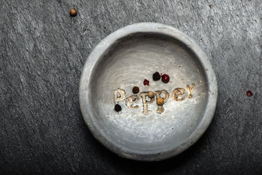 Black pepper in small bowl for spices on dark background. Red, green and black pepper grains close-up and natural light on it. Dark stone background and bowl with text Pepper on the bottom.