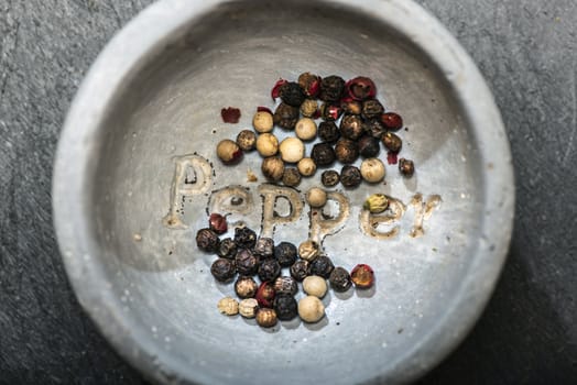 Black pepper in small bowl for spices on dark background. Red, green and black pepper grains close-up and natural light on it. Dark stone background and bowl with text Pepper on the bottom.