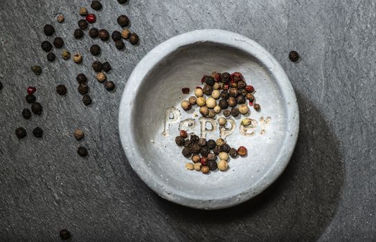 Black pepper in small bowl for spices on dark background. Red, green and black pepper grains close-up and natural light on it. Dark stone background and bowl with text Pepper on the bottom.