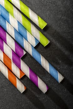 Multicoloured drink straws on dark stone background. Vibrant colours contrast on natural light. Paper made straws. Close-up macro shot.