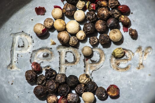 Black pepper in small bowl for spices on dark background. Red, green and black pepper grains close-up and natural light on it. Dark stone background and bowl with text Pepper on the bottom.