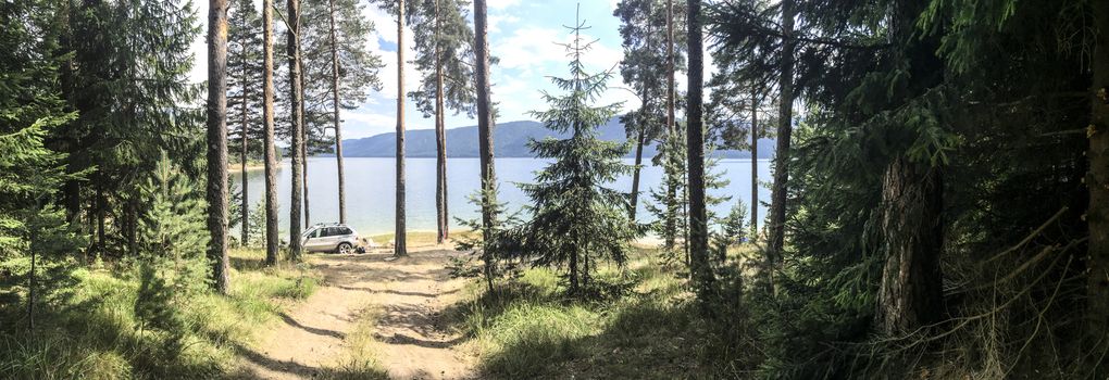 Panoramic image of forest and mountain lake. Tents and car in the wild nature. Sunny day. Many pine trees. Panorama in the forest.