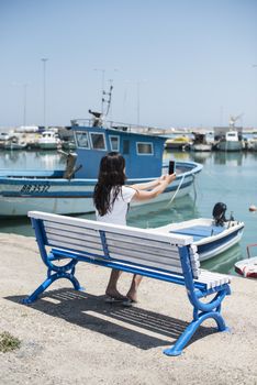 Woman sitting on a bench on the beach in front of boats. 