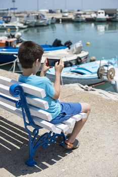 Child sitting on a bench on the beach in front of boats. 